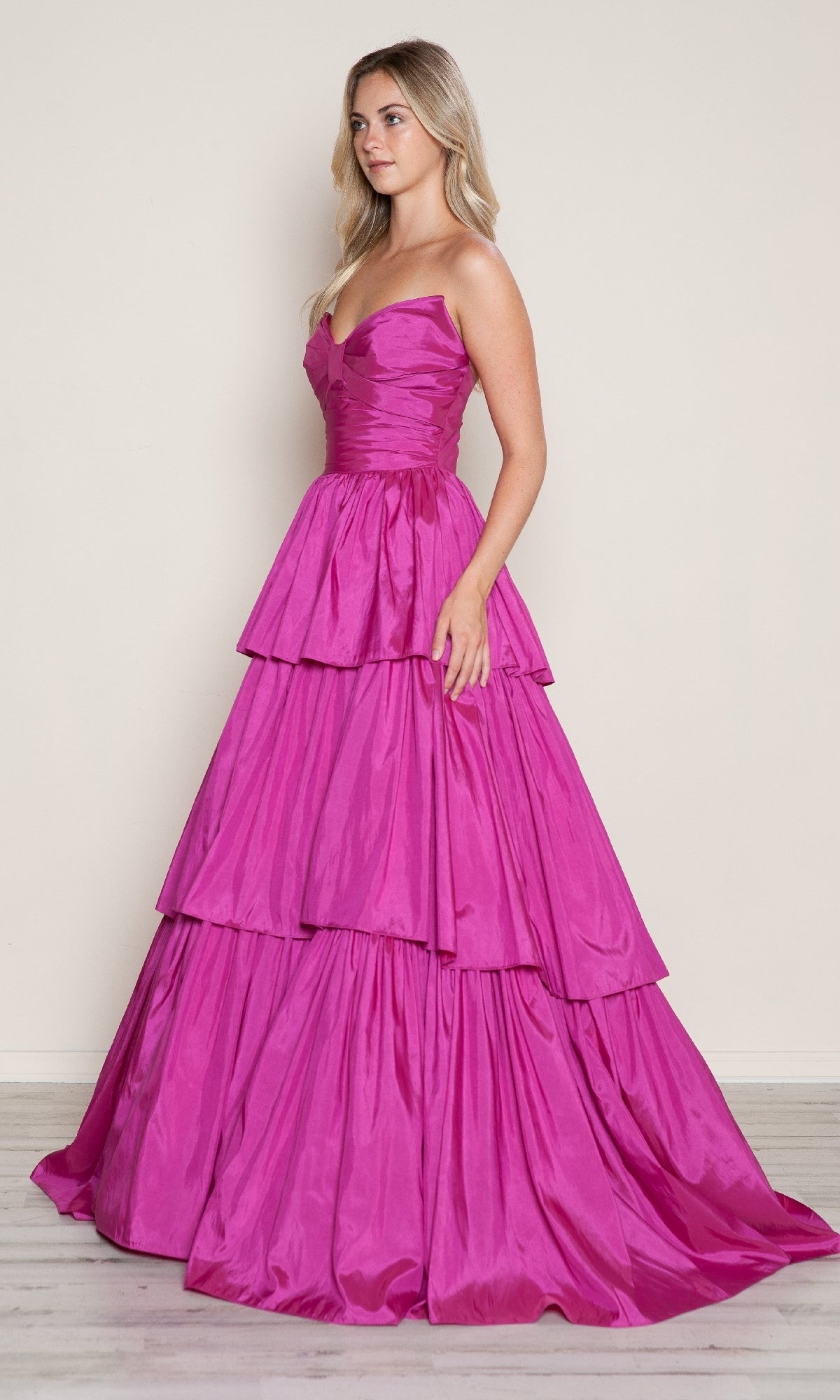 A woman stands in profile wearing the Poly USA 9694 long prom dress—a strapless, bright pink ball gown with a fitted bodice and tiered, floor-length skirt—set against a plain background.